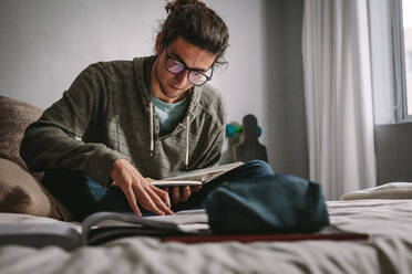 Ein Student sitzt auf einem Bett und studiert konzentriert. Ein junger Mann bereitet sich auf eine Prüfung vor und liest Bücher. - JLPSF24640