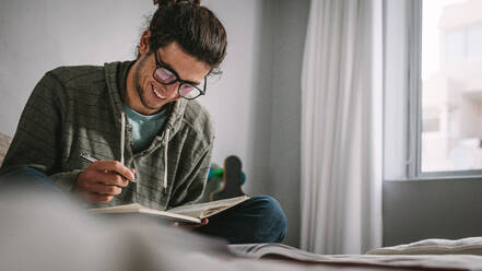 Cheerful young man preparing notes and studying sitting at home beside a window. Student preparing for college exams studying at home sitting on bed. - JLPSF24642