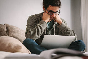 Junger Mann sitzt mit einem Laptop auf dem Bett und sieht besorgt aus. Student studiert ernsthaft für Prüfungen mit Laptop und Büchern auf dem Bett. - JLPSF24644