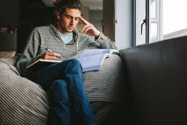 Junger Mann sitzt zu Hause auf einer Couch mit Büchern und denkt nach. Student macht sich Notizen beim konzentrierten Lernen zu Hause. - JLPSF24646