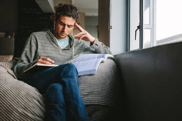 Student sitting at home looking tensed studying seriously. Man sitting on couch beside a window writing in a book. - JLPSF24647