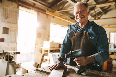 Lächelnder älterer Schreiner an der Werkbank mit einer Bohrmaschine. Glücklicher reifer männlicher Schreiner in seiner Werkstatt. - JLPSF24665