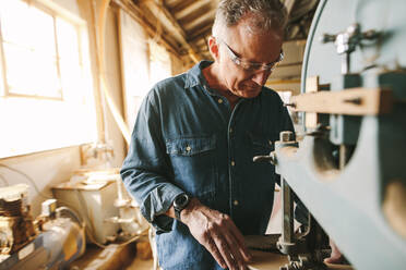 Älterer männlicher Tischler bei der Arbeit in seiner Werkstatt. Ein Tischler schneidet Holz an einer Maschine in einer Tischlerei. - JLPSF24667