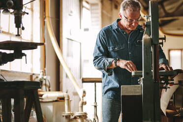 Älterer männlicher Tischler bei der Arbeit an einer Bandsägemaschine in einer Tischlerwerkstatt; älterer Mann schneidet Holz an der Maschine. - JLPSF24670