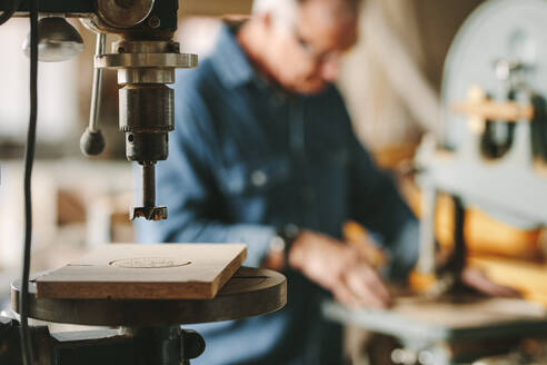 Drilling machine in carpentry workshop with senior carpenter working on band saw in background. - JLPSF24671