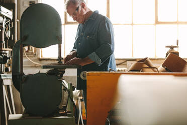 Ein älterer Arbeiter arbeitet an einer Bandsägemaschine in seiner Werkstatt. Ein Tischler schneidet Holz an einer Maschine in einer Tischlerei. - JLPSF24672