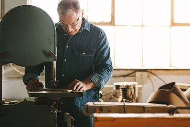 Älterer Arbeiter in der Schreinerei schneidet das Holz mit einer Bandsäge zu. Schreiner in der Schreinerei bei der Vorbereitung von Möbelteilen. - JLPSF24673
