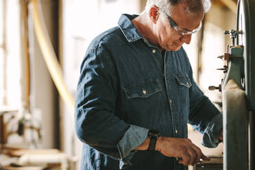 Älterer männlicher Tischler bei der Arbeit in seiner Werkstatt. Ein Tischler schneidet Holz auf einer Bandsäge in der Werkstatt. - JLPSF24674