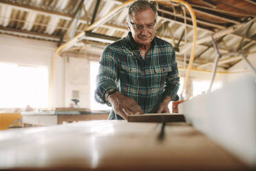 Senior male carpenter working on table saw machine. Man cutting wood planks on table saw machine in carpentry workshop. - JLPSF24677