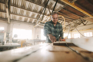 Der leitende Tischler arbeitet an der Maschine für die Herstellung von Holzprodukten. Der Tischler schneidet das Holz in verschiedenen Konfigurationen auf der Kreissägemaschine. - JLPSF24679