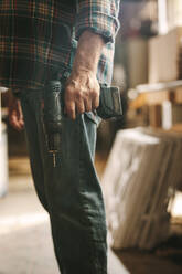 Mittelteil eines Tischlers mit einer Handbohrmaschine in einer Tischlerei. Mann mit Bohrmaschine in der Hand in einer Werkstatt. - JLPSF24685