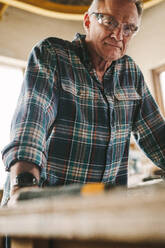 Älterer Mann mit Schutzbrille, der an einer Werkbank steht und in die Kamera schaut. Älterer Zimmermann in seiner Schreinerei. - JLPSF24694
