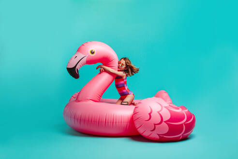 Mädchen in Badekleidung sitzt auf einem riesigen aufblasbaren rosa Flamingo. Kleines Mädchen im Badeanzug auf einer aufblasbaren Spielzeugmatratze vor blauem Hintergrund. - JLPSF24708