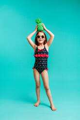 Stylish girl child in swimwear holding a green coloured pineapple on her head. Girl wearing swimwear and sunglasses over blue background. - JLPSF24711