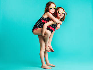Girl carrying her friend on her back. Small twin girls in swimwear piggybacking on blue background. - JLPSF24713