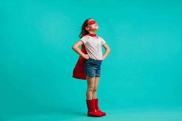Mädchen mit roten Gummistiefeln, Umhang und Augenmaske steht mit den Händen in den Hüften in einem Studio und schaut weg auf blauem Hintergrund. - JLPSF24720