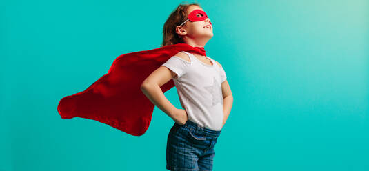 Mädchen in rotem Superheldenkostüm steht mit den Händen auf den Hüften im Studio. Mädchen in rotem Umhang und Maske schaut weg auf blauem Hintergrund. - JLPSF24721
