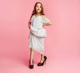 Girl wearing large dress and high heels shoes pretending to be a grown up woman. Girl child posing in oversized clothes on pink background. - JLPSF24735