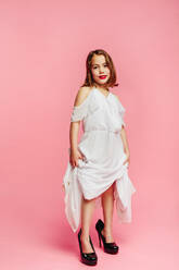 Full length of girl wearing mothers high heel shoes and white dress pretending she's a grown up lady. Caucasian girl child posing on large dress and high heel shoes on pink background. - JLPSF24737