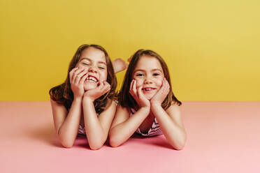 Zwillingsmädchen, die mit den Händen auf dem Kinn auf dem rosa Boden liegen und in die Kamera schauen, auf gelbem Hintergrund. - JLPSF24740