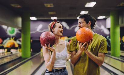 Fröhliches junges Paar auf der Bowlingbahn mit den Kugeln, die sich gegenseitig anschauen. Junger Mann und Frau stehen in einem Bowlingclub und halten Kugeln. - JLPSF24744