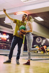 Lächelndes Paar mit Bowlingkugeln, das ein Selbstporträt mit einem Smartphone in einem Club macht. Ein junger Mann und eine Frau machen ein Selfie von sich selbst in einer Bowling-Arena. - JLPSF24747