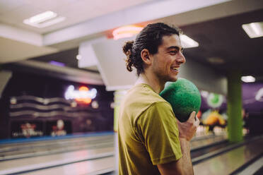 Stilvoller junger Mann auf einer Bowlingbahn mit einer Kugel in der Hand. Kaukasischer Mann mit Kugel im Bowlingclub. - JLPSF24750