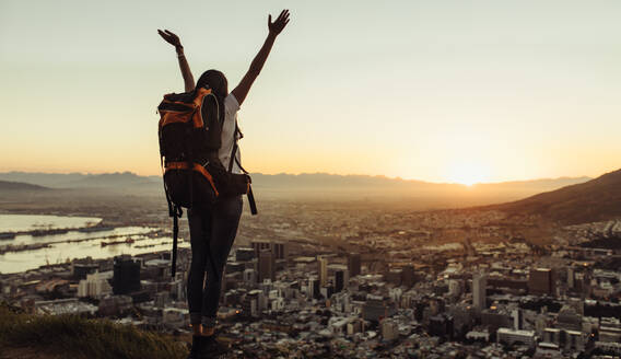 Rückansicht einer weiblichen Reisenden mit erhobenen Händen, die auf der Spitze eines Hügels steht und auf die Stadt mit Sonnenuntergangshimmel blickt. Frau mit Rucksack, die die Aussicht auf die Stadt von der Spitze eines Berges genießt. - JLPSF24774