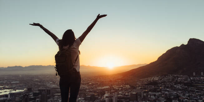 Frau Tourist mit Rucksack genießt große Aussicht stehend auf dem Hügel. Weibliche Reisende stehend mit ihren Händen erhoben Blick auf die Stadt bei Sonnenuntergang. - JLPSF24775