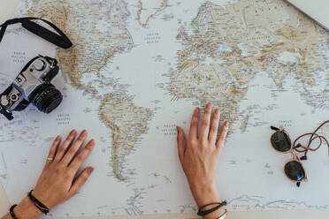Persönliche Perspektive einer Touristin, die eine Weltkarte betrachtet, um ihren Urlaub zu planen, mit einer Kamera und einer Sonnenbrille in der Hand. - JLPSF24785