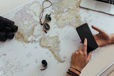 Frau Hand mit Smartphone mit einer Weltkarte auf dem Tisch. Point of view der weiblichen Tourist Planung Urlaub mit Reise-Accessoires um. - JLPSF24786