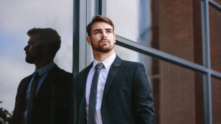 Confident businessman standing by a glass wall looking away outdoors. Man with beard wearing business suit standing outside and looking at copy space thinking. - JLPSF24790