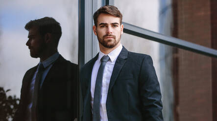 Portrait of handsome young businessman standing by office building and staring at camera. Man in business suit looking at camera while standing outside. - JLPSF24791