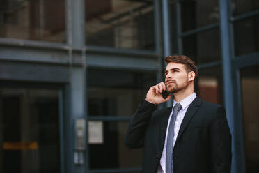 Businessman walking out of office and talking over cell phone. Businessman in formal clothes walking outdoors using mobile phone. - JLPSF24794