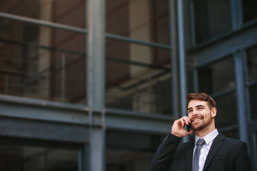 Positive young businessman making a phone call outdoors with office building in background. Business professional talking on cell phone with lot of copy space. - JLPSF24796