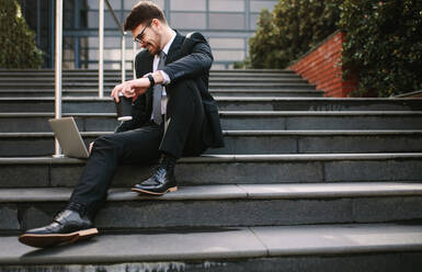 Smiling young business man sitting on steps with laptop and coffee. Relaxed entrepreneur looking at laptop and smiling while sitting on steps outside office. - JLPSF24803