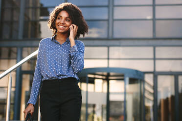 Smiling businesswoman talking over mobile phone while walking outside office building. Woman in formalwear walking outdoors and talking on phone. - JLPSF24805