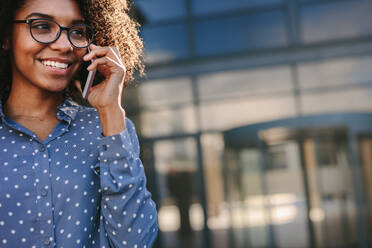 Afrikanische Frau im Hemd spricht am Telefon und lächelt im Freien. Nahaufnahme einer glücklichen jungen Frau, die einen Anruf tätigt, während sie im Freien mit Kopierraum steht. - JLPSF24809