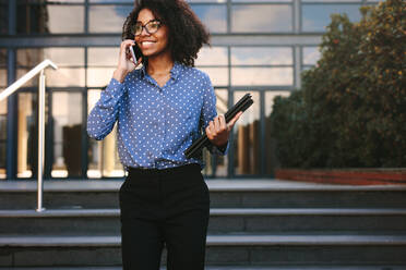Glückliche junge Frau in formeller Kleidung, die eine Akte hält und ein Telefongespräch mit einem Bürogebäude im Hintergrund führt. - JLPSF24810