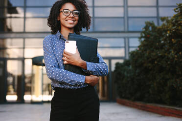 Confident woman in formal clothes holding a smartphone and file looking away smiling. Female executive standing outside office building. - JLPSF24812