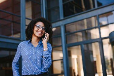 African woman formal clothes talking on cell phone and looking away smiling. Businesswoman standing outside office building talking over mobile phone. - JLPSF24818