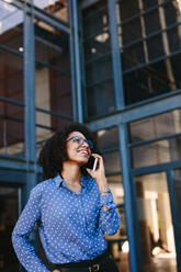 Young female executive standing outside office building talking on cell phone. African businesswoman making a phone call while standing against a corporate office building. - JLPSF24819