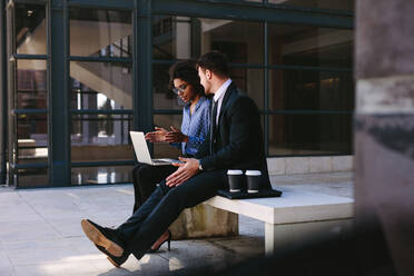 Two business people sitting on a bench in modern office building with a laptop. Woman with laptop discussing business with male colleague. - JLPSF24825