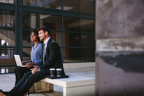 Geschäftskollegen sitzen auf einer Bank in der Bürohalle mit Laptop und schauen lächelnd weg. Geschäftsmann und Frau entspannen sich im Büroflur mit Kaffeetassen an der Seite. - JLPSF24826