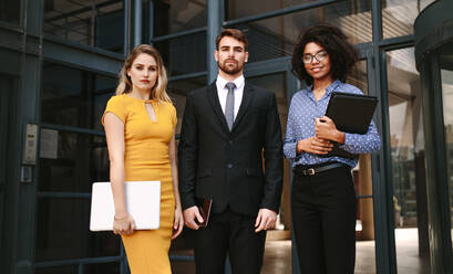 Portrait of three business people standing together in front of office building. Group of diverse business professionals looking at camera confidently. - JLPSF24832