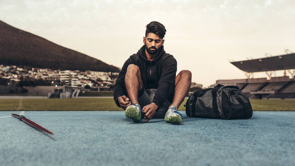 Sportler, der sich in einem Leichtathletik-Stadion mit Schuhen auf das Training vorbereitet; Mann, der auf der Bahn sitzend seine Schuhe zubindet, mit einem Speer und einer Tasche an seiner Seite. - JLPSF24870