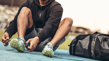 Sportler mit Sportschuhen, der sich auf das Training vorbereitet. Ausschnitt eines Läufers, der auf einer Laufbahn sitzt und die Schnürsenkel bindet, mit einer Tasche an seiner Seite. - JLPSF24871
