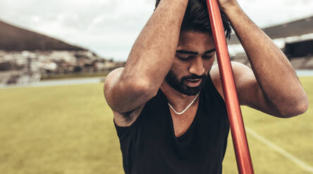 Ein müder Athlet steht auf einem Leichtathletikplatz und hält einen Speer. Ein Athlet entspannt sich während seines Speerwurf-Trainings und hält mit geschlossenen Augen einen Speer. - JLPSF24873