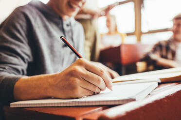 Nahaufnahme eines männlichen Studenten, der sich während einer Vorlesung am Schreibtisch sitzend Notizen macht; der Fokus liegt auf der Hand, die mit einem Bleistift in ein Notizbuch schreibt. - JLPSF24892