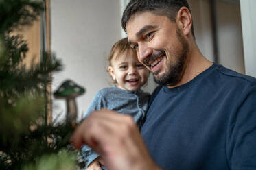 Glücklicher Vater trägt seinen Sohn und schmückt den Weihnachtsbaum zu Hause - ANAF00330